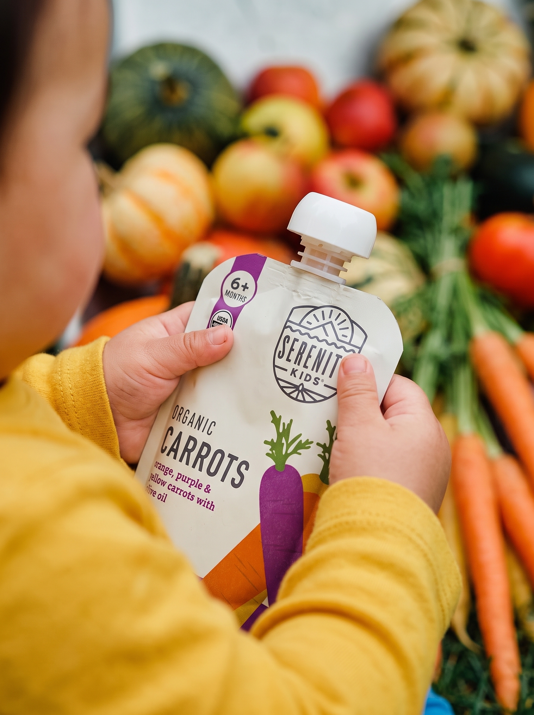A close view of small hands holding a Serenity Kids carrots pouch among fall produce