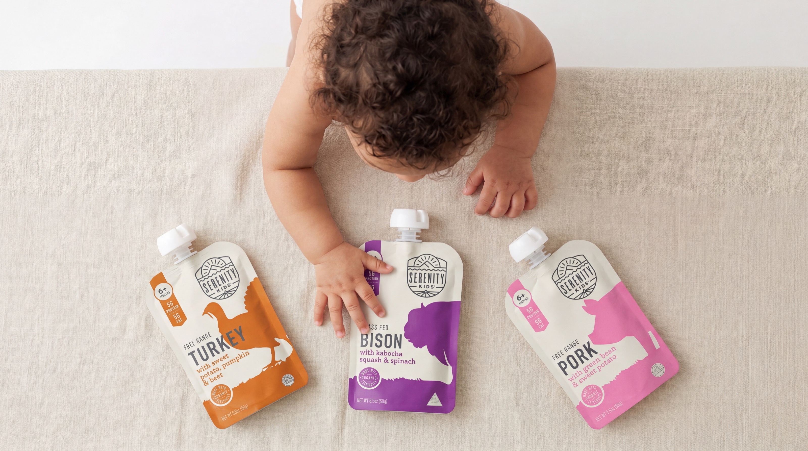 A toddler reaching across a cream sofa toward three Serenity Kids food pouches